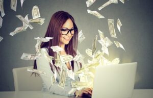 A young woman trading stocks at her laptop while money rains down. She may be looking for the best stocks to buy for beginners.